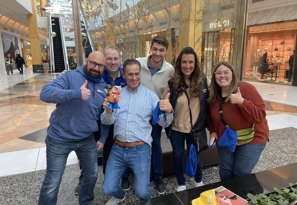 Group of six participants smiling and giving thumbs up during a team building event in a shopping mall, holding a Mr. Potato Head toy.
