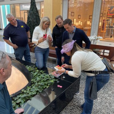 Team building participants gather around a table in a mall as one blindfolded member engages in a challenge involving assembling a toy. thumbnail