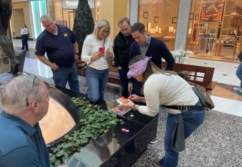 Team building participants gather around a table in a mall as one blindfolded member engages in a challenge involving assembling a toy.
