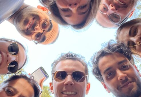 A group of smiling team members huddled in a circle, looking down at the camera during a fun and engaging outdoor team building activity.