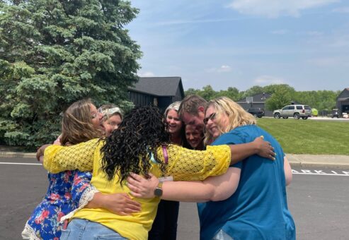A group of six teammates sharing a heartfelt group hug outdoors during a team building event, standing on a paved road with greenery and trees in the background.