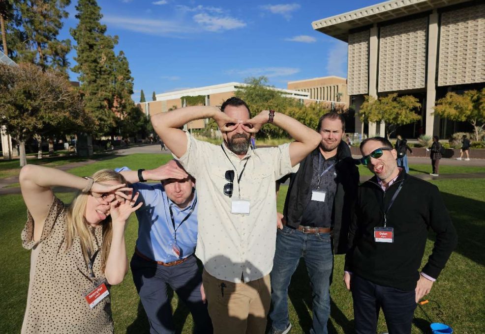 Team building participants strike playful poses while standing on a grassy area outside a building.