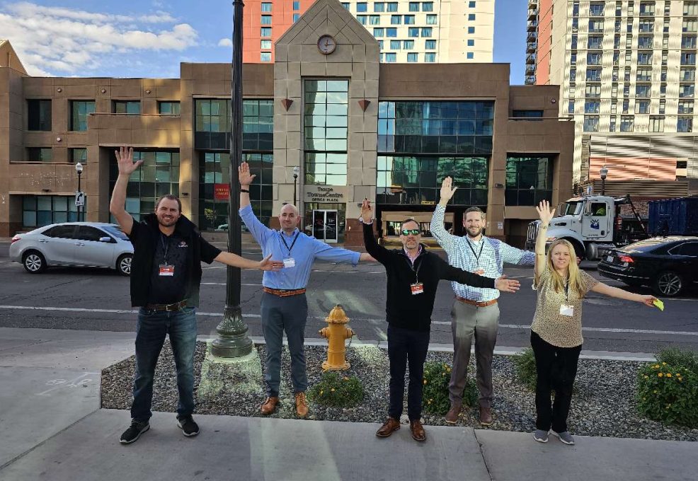 Five team members standing outside, smiling and raising their arms enthusiastically as part of a team building activity during a scavenger hunt, with an urban backdrop of buildings behind them.