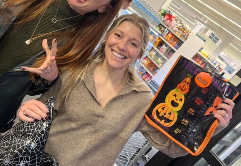 Two women are standing in what appears to be a grocery store, smiling and posing for the camera. The woman on the left has long red hair, is wearing a green sweater, and is making a playful spooky face with her hands raised as if pretending to be a monster. The woman on the right is smiling brightly and holding up a Halloween-themed reusable shopping bag with a jack-o'-lantern design. The scene is part of a Halloween-themed team building challenge.