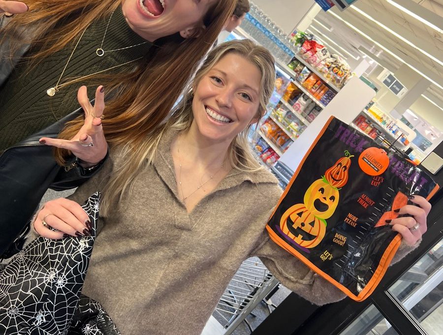 Two women are standing in what appears to be a grocery store, smiling and posing for the camera. The woman on the left has long red hair, is wearing a green sweater, and is making a playful spooky face with her hands raised as if pretending to be a monster. The woman on the right is smiling brightly and holding up a Halloween-themed reusable shopping bag with a jack-o'-lantern design. The scene is part of a Halloween-themed team building challenge.