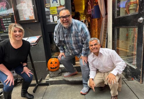 Three people are crouching and smiling outside a building with a carved pumpkin placed on the steps beside them. The person on the left is a woman with blonde hair, wearing a black top and jeans. In the middle is a man wearing glasses, a plaid shirt, and gray pants. The person on the right is another man dressed in a white shirt and khaki pants. The group is engaging in a Halloween-themed team building activity.