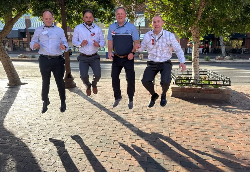 Four men participating in a team building activity captured mid-air while jumping on a sunlit brick pathway with trees and buildings in the background.