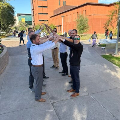 A group of men forming an arch with their arms during a team building exercise outdoors, with a red brick building in the background. thumbnail