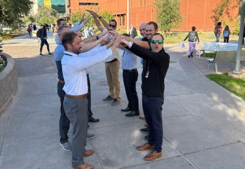 A group of men forming an arch with their arms during a team building exercise outdoors, with a red brick building in the background.