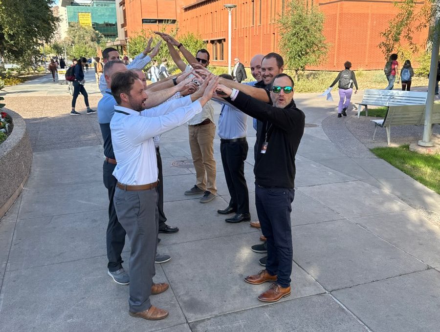 A group of men forming an arch with their arms during a team building exercise outdoors, with a red brick building in the background.