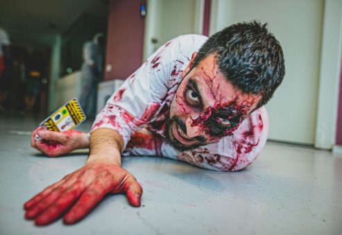 A person dressed as a bloodied zombie crawls on the floor with a fierce expression. The scene is part of a zombie-themed team building event, with realistic makeup and blood effects creating a spooky atmosphere.