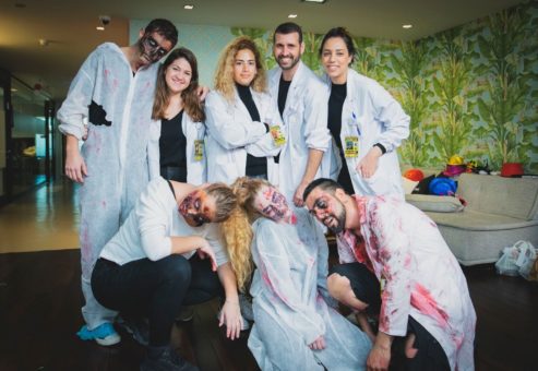 A group of people dressed in white lab coats and zombie makeup, posing together indoors. Some are wearing realistic zombie face paint and bloody costumes, while others are in scientist attire, creating a dynamic contrast between survivors and the undead in a zombie-themed team building event.