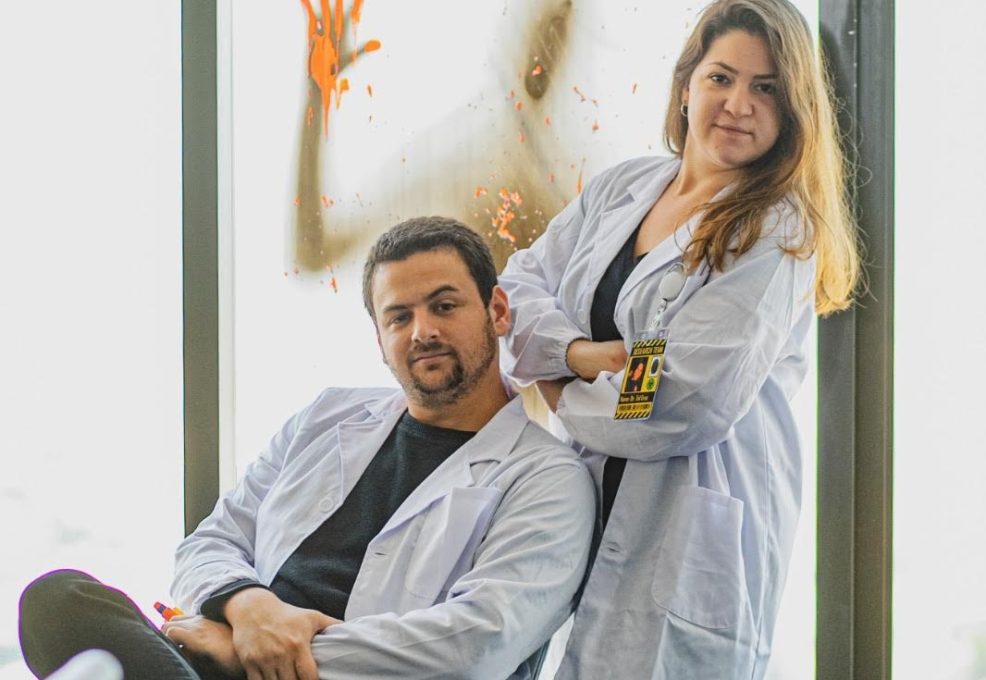 Two individuals dressed as scientists, wearing white lab coats, pose in front of a window with 'HELP' written in red on the glass. Behind the glass, a shadowy figure with a hand pressed against the window creates an eerie, zombie-like atmosphere. The scene is part of a zombie-themed team building event.