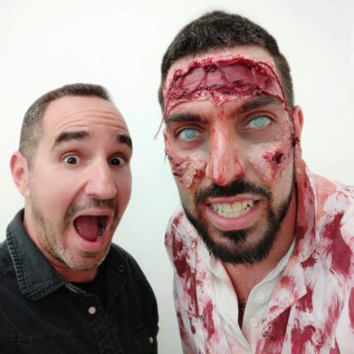 A man excitedly poses next to a participant dressed as a zombie, complete with realistic prosthetic makeup featuring bloody wounds, decaying skin, and white contact lenses, creating an eerie and convincing undead appearance during a zombie-themed team building event. thumbnail