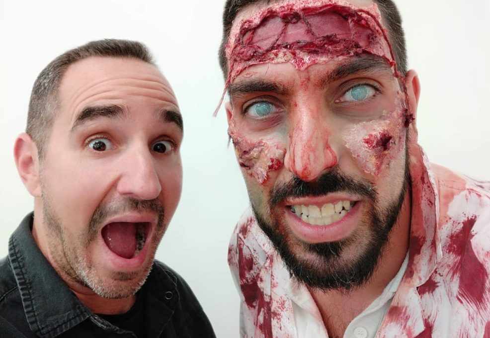 A man excitedly poses next to a participant dressed as a zombie, complete with realistic prosthetic makeup featuring bloody wounds, decaying skin, and white contact lenses, creating an eerie and convincing undead appearance during a zombie-themed team building event.