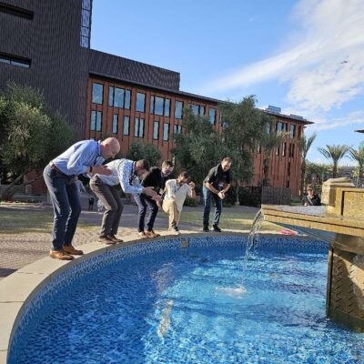 Group of five team building participants crouched near a water fountain, preparing to take action during an outdoor team building activity. thumbnail
