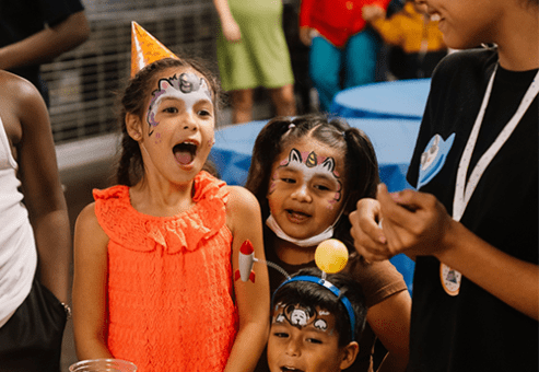 Three children with painted faces are excitedly smiling and laughing at a party. The child in the front is wearing an orange party hat, and they are surrounded by other attendees and decorations, creating a festive atmosphere.
