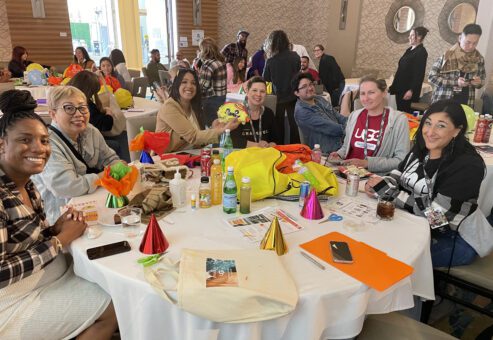 A group of people seated around a table, smiling and interacting, with colorful party hats, birthday-themed decorations, and supplies spread out, engaging in a team building activity for a charitable cause.