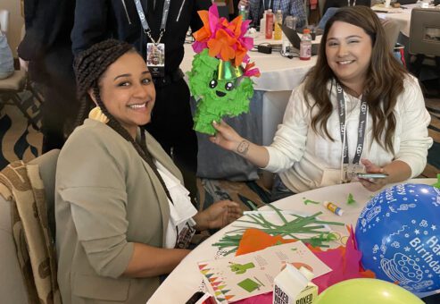 Two women smile while holding up a green cactus-themed piñata decorated with colorful flowers, sitting at a table filled with birthday craft supplies and party decorations. Balloons add to the celebratory mood of this charitable team building program.
