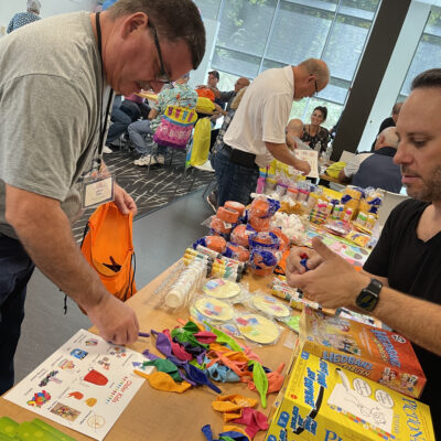 Two men are selecting colorful balloons, party supplies, and games from a table filled with items like 