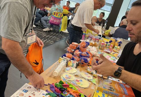 Two men are selecting colorful balloons, party supplies, and games from a table filled with items like 
