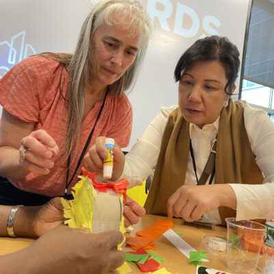 Two women are working together, focused on crafting a colorful project using glue and paper. They are engaged in a team building activity at a table with various supplies to create a piñata as part of a birthday kit donation. thumbnail