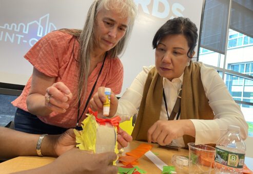 Two women are working together, focused on crafting a colorful project using glue and paper. They are engaged in a team building activity at a table with various supplies to create a piñata as part of a birthday kit donation.