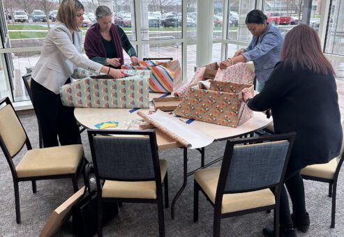 Four people are gathered around a table wrapping large boxes with colorful gift paper, creating a festive atmosphere as they prepare for birthday kits to donate as part of a team building event.