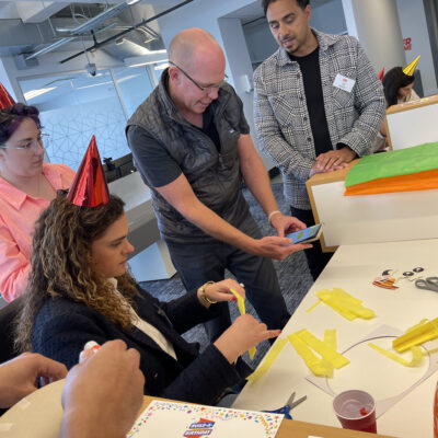 A group of people wearing party hats are engaged in a crafting activity. One person is seated, cutting yellow paper strips, while others stand around, observing and discussing the team building activity. thumbnail