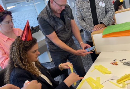 A group of people wearing party hats are engaged in a crafting activity. One person is seated, cutting yellow paper strips, while others stand around, observing and discussing the team building activity.