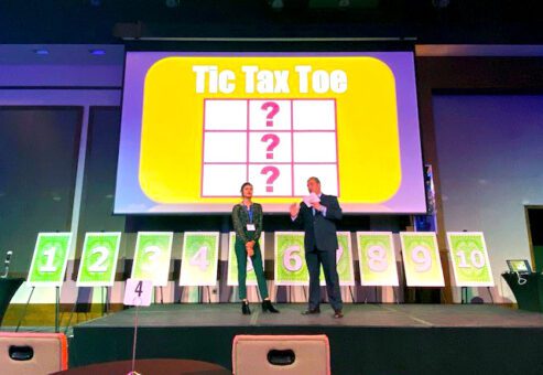 Two hosts stand on stage during the Play it Forward game show event, with a large screen behind them displaying the title 'Tic Tax Toe.' The event is a charitable team building activity where participants compete in pricing and trivia games to win for charity.