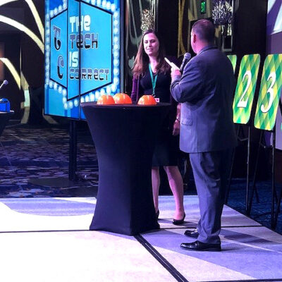 A host engages a contestant in the Play it Forward game show event on stage, with a screen in the background displaying the phrase 'The Tech is Correct.' The stage is set for a trivia challenge, and the contestant stands next to a table with orange buttons used in the game. Numbered boards are visible on the side, representing various challenges in this charitable team building activity. thumbnail