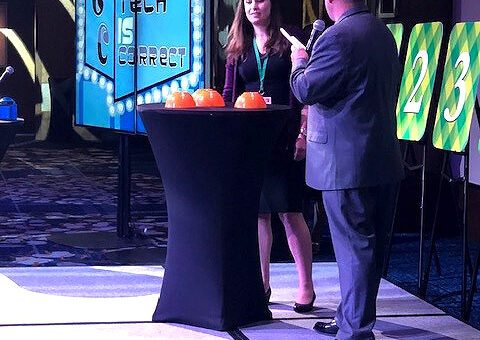 A host engages a contestant in the Play it Forward game show event on stage, with a screen in the background displaying the phrase 'The Tech is Correct.' The stage is set for a trivia challenge, and the contestant stands next to a table with orange buttons used in the game. Numbered boards are visible on the side, representing various challenges in this charitable team building activity.