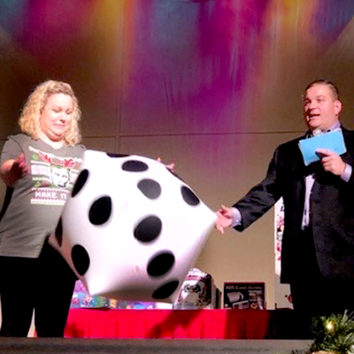 A contestant rolls a large inflatable die on stage during the Play it Forward charity team building game show event, while the host watches and holds game cards. thumbnail