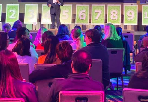 A host stands on stage in front of a row of numbered boards during the Play it Forward charity game show. The audience watches attentively as the host explains the next challenge in this interactive team building event, where all games and activities are designed to raise funds and donations for charity.
