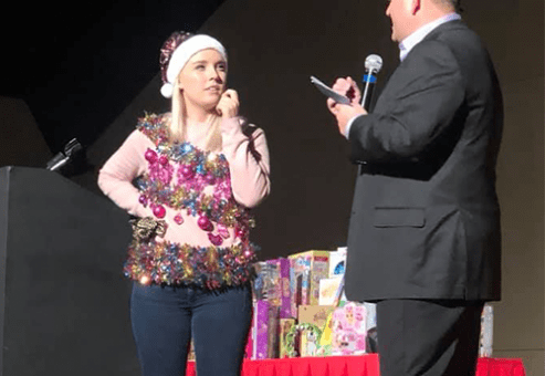A contestant dressed in festive attire stands on stage with a host during the Play it Forward charitable team building game show. The host asks the contestant a question as they participate in one of the interactive challenges.