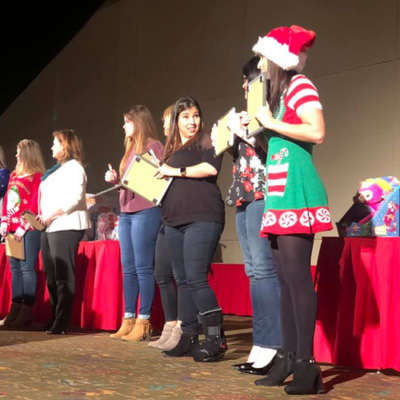 Participants stand in a row on stage during the Play it Forward charity game show, with some wearing festive holiday attire. Each holds a clipboard, preparing for a fun challenge as part of the team building event. thumbnail