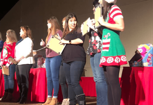 Participants stand in a row on stage during the Play it Forward charity game show, with some wearing festive holiday attire. Each holds a clipboard, preparing for a fun challenge as part of the team building event.