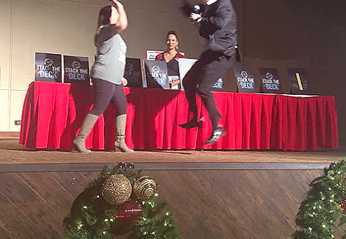 A contestant and host enthusiastically jump and high-five on stage during the Play it Forward charity game show. The event features exciting games like 'Stack the Deck,' with prizes and challenges, all aimed at raising donations for charity. The festive decorations add to the lively and energetic atmosphere of this team building activity.