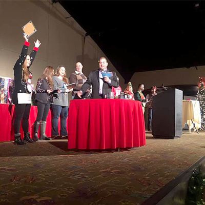 Contestants line up on stage during the Play it Forward charity game show, with one participant raising their hand excitedly as they prepare for the next challenge. The host stands at the center, holding game cards. This team building event involves fun, interactive games, and raises donations for charity. thumbnail