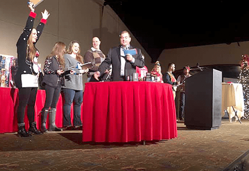 Contestants line up on stage during the Play it Forward charity game show, with one participant raising their hand excitedly as they prepare for the next challenge. The host stands at the center, holding game cards. This team building event involves fun, interactive games, and raises donations for charity.