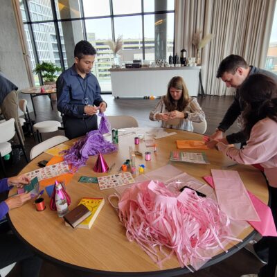 A group of people is gathered around a table, engaged in a crafting activity. The table is covered with materials like colorful paper, party hats, stickers, and pink streamers as they work on assembling birthday kits to be donated as a result of this charitable team building program. thumbnail