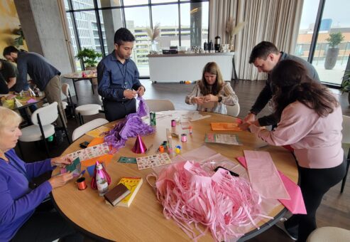 A group of people is gathered around a table, engaged in a crafting activity. The table is covered with materials like colorful paper, party hats, stickers, and pink streamers as they work on assembling birthday kits to be donated as a result of this charitable team building program.