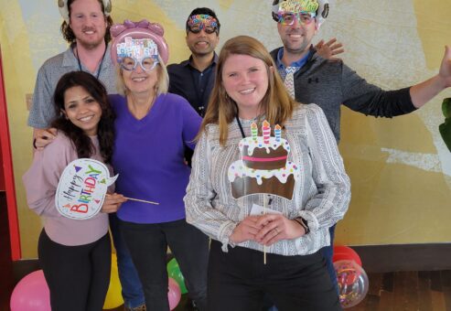 A group of six people poses happily in front of a 'Happy Birthday' banner. They're holding birthday-themed props, surrounded by colorful balloons, and wearing fun hats and party accessories during a team building event.