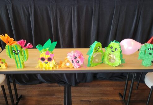 A row of colorful and playful piñatas, including cacti and other fun shapes, is displayed on a table. Some are wearing party hats, sunglasses, or decorated with balloons, bringing a festive vibe to the birthday kits that are going to be donated as a result of this team building activity.