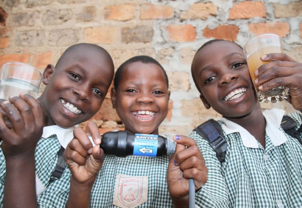 Three smiling children hold clean water glasses, showcasing the impact of Clean Water Connection's filtration systems. These systems provide access to safe drinking water in communities worldwide, helping empower people through clean water initiatives.