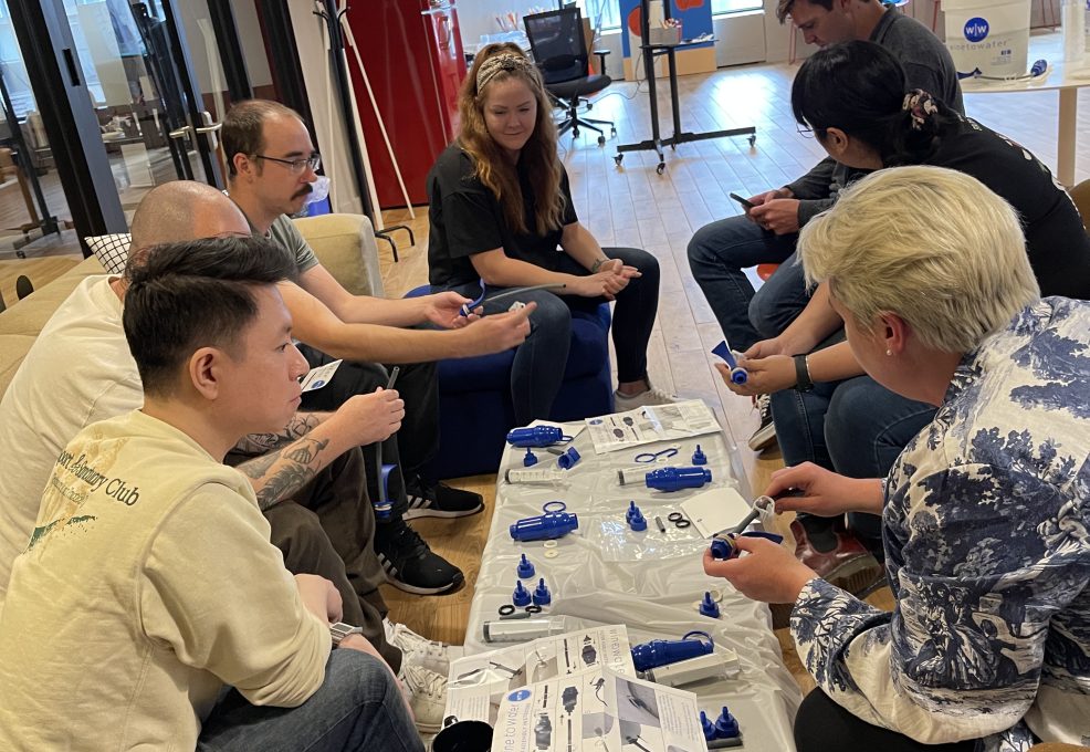 A diverse team of adults engaging in a hands-on clean water filter assembly as part of the Clean Water Connection event. The group works together in a collaborative environment, assembling water filtration kits while learning about the global water crisis. The interactive team building activity fosters community engagement.
