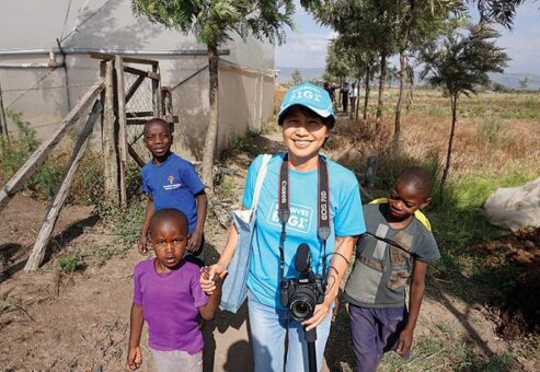A woman wearing a blue B1G1 cap smiles warmly as she stands outside with three young children in a rural area. She is holding a camera, and the children, two boys and one girl, appear happy and curious. This photo captures a moment of connection and joy as donations are being delivered to support the community.