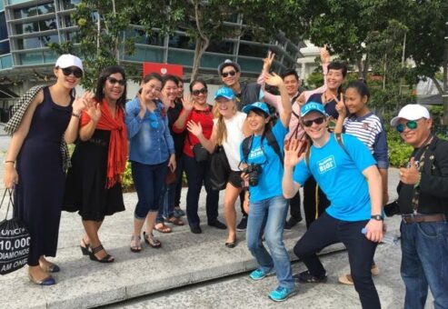 A group of cheerful participants posing for a photo outdoors during a charity team building challenge. They are smiling and waving. This image captures the joy and camaraderie of teams engaging in the Go Give charity challenge, where participants complete GPS-triggered tasks to positively impact the community.