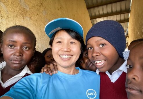 A woman in a blue B1G1 shirt and cap takes a selfie with a group of school children in red uniforms, who gather around her with bright smiles. This image reflects the positive impact of the donations on the local community.